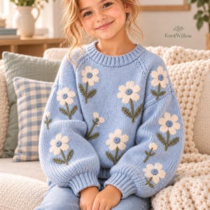 Puede incluir: Un suéter de punto de manga larga en color azul claro con un patrón repetitivo de flores blancas con tallos y hojas verdes. El suéter tiene un cuello redondo acanalado y puños acanalados. Se incluye un pasador de pelo de flor azul.