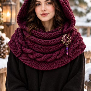 May include: A deep burgundy chunky knit hooded cowl featuring a thick cable knit border and a gold-toned crescent moon brooch with leaf accents and dangling purple beads, worn over a solid black long-sleeved garment.