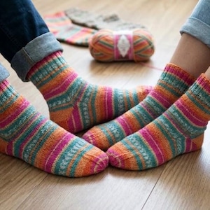 Puede incluir: Dos pares de calcetines tejidos a mano con un vibrante patrón de rayas multicolor en tonos rosa, naranja, turquesa y amarillo. Los calcetines tienen un puño acanalado y un diseño de punto texturizado. Un ovillo de lana a juego está al fondo.