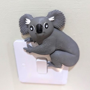 Puede incluir: Una cubierta de interruptor de luz con forma de koala gris con acabado texturizado, grandes ojos negros con reflejos blancos, nariz negra y mechones de pelo blanco en las orejas. El koala está posicionado como si se aferrara a la placa blanca.