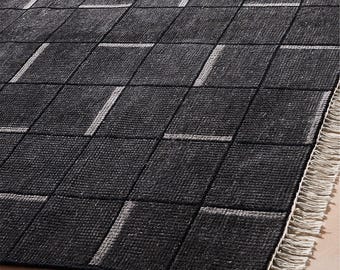 Puede incluir: Una alfombra rectangular con un patrón de cuadrícula gris carbón oscuro con acentos rectangulares alternos en gris claro horizontales y verticales dentro de los cuadrados. La alfombra tiene una superficie tejida texturizada y un borde con flecos color hueso.