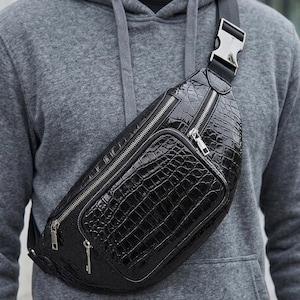Könnte beinhalten: Eine schwarze Bauchtasche mit glänzender Krokodilleder-Optik, ausgestattet mit einem Hauptfach mit Reißverschluss und einer kleineren Fronttasche mit Reißverschluss. Die Tasche hat einen verstellbaren schwarzen Gurt mit silberner Metallschnalle.