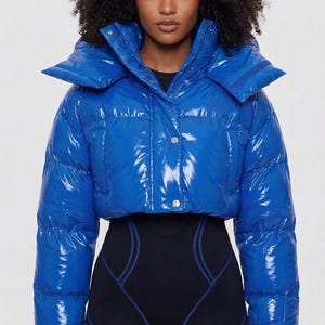 Puede incluir: Una chaqueta acolchada corta de color azul real brillante con cuello ancho, mangas largas y cierre frontal con botones a presión. La chaqueta presenta una construcción acolchada con un acabado brillante, sobre un body negro con costuras azules.