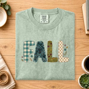 以下が含まれることがあります： セージグリーンの半袖Tシャツで、大きなブロック体で「FALL」という文字がプリントされています。各文字には、ティール色のバッファローチェック、ティール色のヒョウ柄、オリーブ色の花柄、小さな花のアクセントが付いたタン色の水玉模様という異なるパターンが施されています。