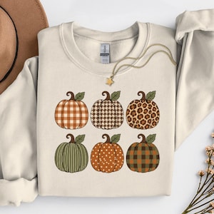 May include: A cream-coloured crewneck sweatshirt featuring a graphic print of six pumpkins in two rows. The pumpkins display various patterns including orange plaid, brown houndstooth, leopard print, green stripes, orange polka dots, and green plaid.