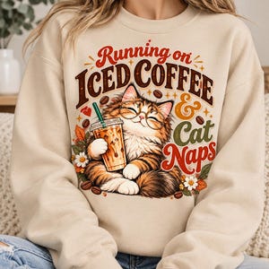 May include: A beige crewneck sweatshirt featuring a graphic of a fluffy cat wearing round glasses and holding an iced coffee. The text reads Running on ICED COFFEE & Cat Naps, surrounded by coffee beans, small flowers, and autumn leaves.