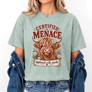 Op de afbeelding: Een licht saliegroen t-shirt met een afbeelding van een bruine Schotse Hooglander met een madeliefje op zijn kop die zijn tong uitsteekt. De tekst "CERTIFIED MENACE" staat erboven en "approach with snacks" op een houten bord onder de koe.