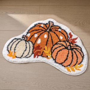 Op de afbeelding: Een onregelmatig gevormd vloerkleed met een witte achtergrond en een getuft ontwerp van drie pompoenen in oranje, crème en lichtoranje. De pompoenen zijn geaccentueerd met kleine witte stippen, donkerbruine omlijningen en verspreide gele en rode herfstbladeren.