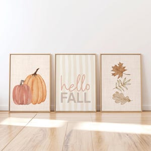 May include: A set of three framed wall art prints. The left print features two watercolor pumpkins in shades of orange and peach. The center print displays vertical stripes with the text hello FALL. The right print shows three brown autumn leaves.