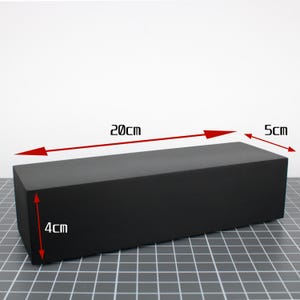 May include: A solid black rectangular block with a matte finish. Red arrows and text indicate the dimensions as 20 cm in length, 5 cm in width, and 4 cm in height. The object is positioned on a surface with a white grid pattern.