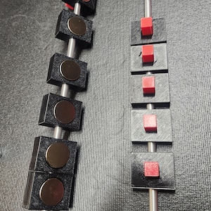 May include: Two vertical metal rods each featuring five black square blocks. The left rod has blocks with circular metallic centers, while the right rod has blocks with small red square protrusions on their front surfaces.