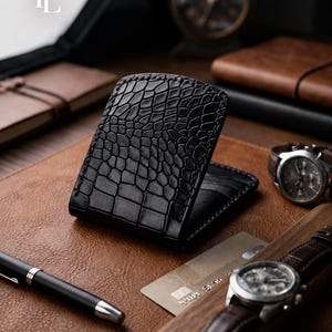 Pode incluir: Uma carteira bifold de couro preto com um padrão de pele de crocodilo texturizado no exterior. A carteira é construída com costuras visíveis ao longo das bordas e é mostrada em uma posição entreaberta, revelando compartimentos para cartões.
