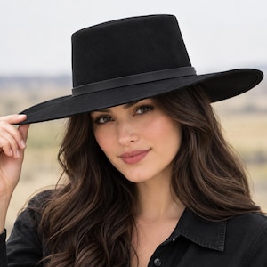 May include: A solid black wide-brim hat with a flat crown and a thin, matching black band encircling the base of the crown. The hat features a structured, stiff brim that extends outward, providing a clean, uniform silhouette in a matte finish.