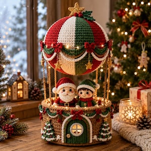 Op de afbeelding: Een gehaakte heteluchtballon-decoratie met rode, witte en groene panelen en een ster als top. De mand is bruin met een groene deur, twee kleine kerstbomen en twee gehaakte figuren van de Kerstman en een elfje in de mand.