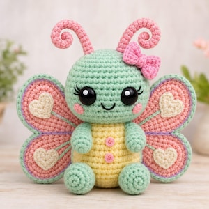 May include: A crocheted butterfly plush toy with a mint green head and body, a yellow belly, and pink wings featuring cream-colored heart appliques. The toy has large black eyes, pink cheeks, two curly antennae, a small pink bow, and two pink buttons.