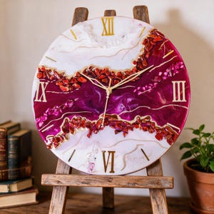 Può includere: Un orologio da parete rotondo con un design in resina bianco e magenta intenso con numeri romani dorati. Il centro è accentuato da pietre frantumate rosse e rosa incastonate, creando un effetto geode testurizzato. Lancette dorate indicano l'ora.