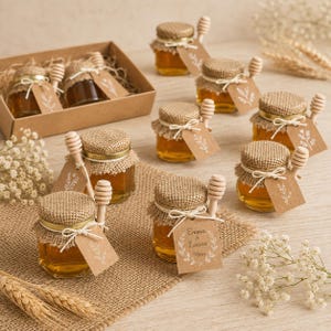 Pode incluir: Pequenos frascos de vidro hexagonais cheios de mel dourado, cada um com uma tampa de tecido de juta, atado com cordão branco e um pau de mel de madeira. Uma etiqueta de papel castanho está presa a cada frasco, algumas com estampas florais.