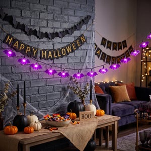 May include: A string of purple bat-shaped LED lights draped across a table decorated for Halloween. The display includes a black banner with the text HAPPY HALLOWEEN, orange pumpkins, black candles, a bowl of candy, and a wooden sign reading BOO.