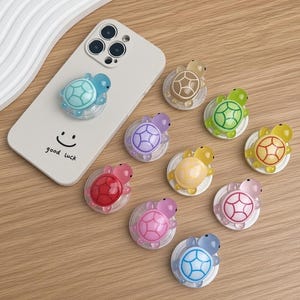 May include: A light-colored smartphone case with a smiley face and the text good luck, featuring a blue turtle-shaped phone grip. Eight additional turtle-shaped phone grips in various translucent colors including beige, green, purple, yellow, red, pink, and blue are arranged nearby.