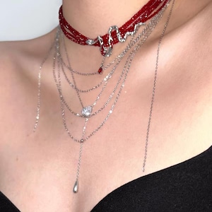 May include: A multi-layered necklace set featuring a deep red beaded choker with a silver-toned snake charm and a dangling red teardrop accent. Below the choker are several delicate silver-toned chains, one with a clear crystal pendant.