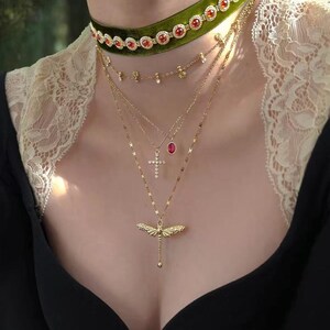 May include: A green velvet choker featuring a row of circular red stones set in gold metal, layered with three gold-tone necklaces. The necklaces include a delicate chain with small charms, a cross pendant, a red oval stone, and a dragonfly charm.