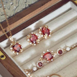 Puede incluir: Un conjunto de joyería en tono dorado que incluye un collar, un par de pendientes y una pulsera. Cada pieza presenta una gran piedra preciosa de color rojo intenso con corte ovalado, rodeada por pequeñas piedras blancas y detalles florales.