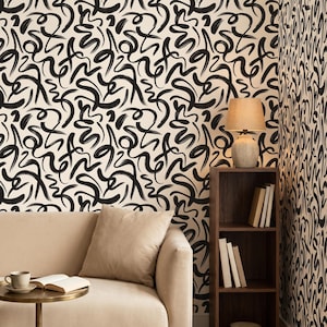 Puede incluir: Papel pintado con un patrón repetitivo de garabatos y bucles negros pintados a mano sobre un fondo crema. El diseño cubre las paredes detrás de un sofá beige, una pequeña mesa redonda con un libro y una taza, y una estantería de madera alta.