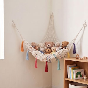 May include: A cream-colored macrame corner hammock mounted to walls with hooks, holding a collection of various plush teddy bears. The hammock features decorative tassels in orange, blue, red, and purple hanging from the bottom edge.