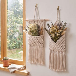 May include: Two off-white macrame wall hangings with pockets holding dried flowers. The left piece features a wooden dowel and fringe, while the right piece has a triangular top and tassel fringe. Both are crafted from knotted cotton cord.