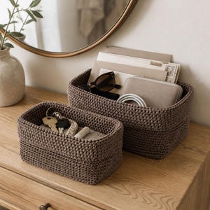 May include: Two rectangular brown woven storage baskets with a thick, textured knit pattern. The larger basket contains envelopes, sunglasses, a beige wallet, and a white cable. The smaller basket holds a set of metal keys and a small white tube.