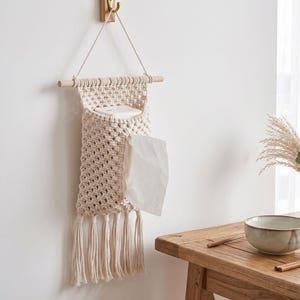 May include: A cream-colored macrame wall hanging tissue holder featuring a woven diamond pattern, a wooden dowel support, and a hanging cord. The holder has a bottom fringe of tassels and contains a white tissue box with a single tissue pulled out.
