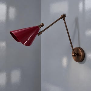 Peut inclure: Une applique murale dotée d'un abat-jour conique rouge foncé fixé à un bras métallique articulé de couleur bronze. Le luminaire comprend une plaque murale circulaire en bronze et une rotule reliant l'abat-jour au bras réglable.