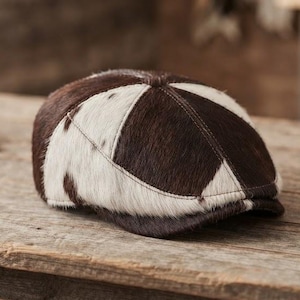 May include: A flat cap constructed from panels of dark brown and white cowhide with a textured, hairy finish. The cap features a rounded crown and a short, stiff brim, with visible stitching connecting the alternating dark and light fur segments.