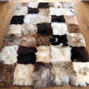 Op de afbeelding: Een rechthoekig vloerkleed opgebouwd uit een patchwork van individuele schapenvachten. Het kleed heeft een meerkleurig palet bestaande uit verschillende tinten wit, crème, beige, bruin, donkerchocolade en zwarte vachtstukken in een patroon.