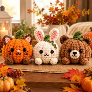 May include: Three crocheted plush figures shaped like pumpkins with animal features. The left is an orange cat, the center is a white rabbit, and the right is a brown bear. Each has black button eyes, embroidered facial details, and green stems.