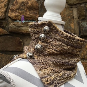 May include: A brown and beige knitted cowl with a cable knit pattern and silver heart-shaped buttons. The cowl is displayed on a white mannequin.