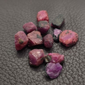 May include: A collection of small, raw, unpolished ruby gemstones in varying shades of deep pink and red. Each stone features an irregular, natural shape with visible dark inclusions and a matte, textured surface across the cluster of crystals.