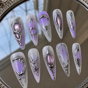 Pode incluir: Um conjunto de dez unhas postiças em formato amendoado com um degradê de branco para roxo. Cada unha é decorada com detalhes metálicos prateados em relevo, incluindo formas de estrelas, luas crescentes e padrões orgânicos fundidos.