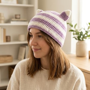 Puede incluir: Un gorro de ganchillo con rayas horizontales en tonos alternos de violeta claro y blanco. El gorro está diseñado con dos orejas puntiagudas similares a las de un gato en la parte superior, hechas con el mismo patrón de hilo rayado.