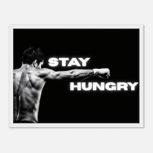 Puede incluir: Una impresión en blanco y negro que muestra a una persona musculosa vista desde atrás, lanzando un puñetazo con la mano vendada. El texto STAY HUNGRY aparece en letras mayúsculas blancas brillantes sobre un fondo negro sólido.