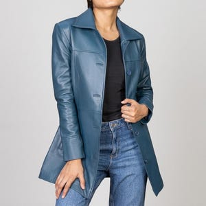 Puede incluir: Una chaqueta de cuero color verde azulado de manga larga con cuello clásico, cierre frontal de botones y costuras estructuradas. La chaqueta se lleva sobre una camiseta negra lisa y vaqueros azules, mostrando un acabado liso y mate.