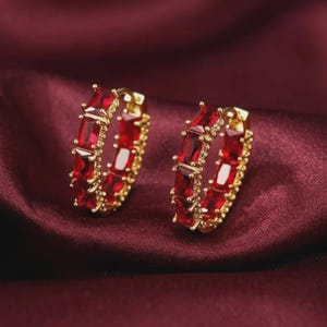 May include: A pair of gold hoop earrings featuring a series of rectangular-cut, deep red gemstones set along the outer edge. The earrings have a polished gold metal finish and a hinged closure mechanism, showcasing a vibrant and symmetrical design.