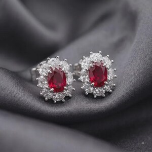 May include: A pair of stud earrings featuring oval-cut deep red rubies as the center stones. Each ruby is surrounded by a halo of twelve round-cut clear white diamonds set in a bright silver-toned metal, creating a floral-inspired cluster design.