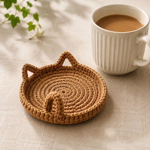 May include: A round, brown crocheted coaster featuring two small, pointed cat-ear shapes along the top edge. The coaster is constructed with a dense, circular stitch pattern and is positioned next to a white, ribbed ceramic mug filled with coffee.