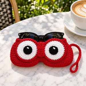 May include: A bright red crochet glasses case featuring two large, circular white and black eye designs on the front. The case has a matching red crochet wrist strap attached to the side and holds a pair of black-framed sunglasses inside.