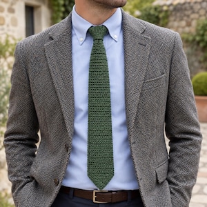 May include: A dark green crochet necktie worn with a light blue button-down shirt and a grey herringbone blazer. The outfit includes a brown leather belt with a gold-toned buckle, creating a textured and layered appearance for formal attire.