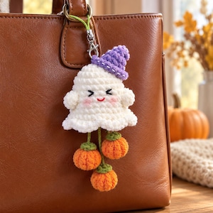 May include: A crocheted ghost keychain featuring a white ghost with a purple hat, pink cheeks, and a smiling face. Three small orange crocheted pumpkins with green stems hang from the bottom of the ghost by green yarn strings, attached to a metal clasp.