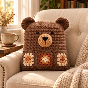 Può includere: Un cuscino all'uncinetto a forma di orso marrone con due piccole orecchie arrotondate, occhi a perla neri e un muso marrone chiaro con un naso ricamato nero. La parte inferiore del cuscino è decorata con tre motivi quadrati all'uncinetto.