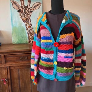 Op de afbeelding: Een gehaakt vest met lange mouwen en horizontale strepen in diverse kleuren, waaronder rood, blauw, roze, groen en geel. Het kledingstuk is voorzien van een fel turquoise rand langs de opening aan de voorzijde en drie grote, ronde knopen.