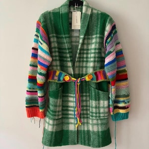 Op de afbeelding: Een groen en wit geruit vest met lange, veelkleurige gestreepte gehaakte mouwen. Het kledingstuk is voorzien van een bijpassende gehaakte riem met twee gele knopen in de taille, twee voorzakken en een sjaalkraag, hangend aan een zwarte kledinghanger.